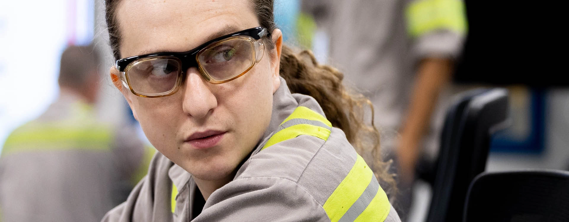 A person wearing safety glasses and a grey work uniform with yellow stripes looks to the side in an industrial setting.