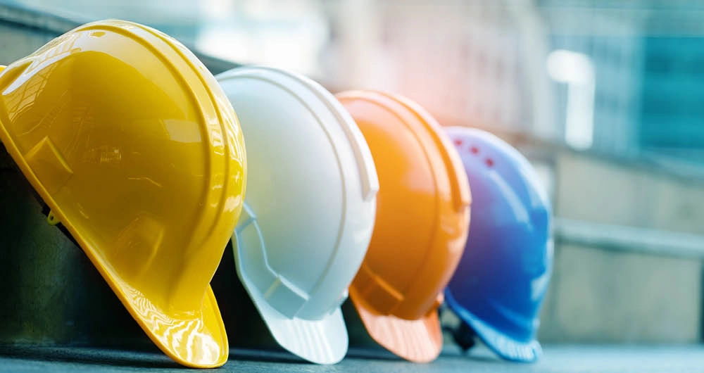 Four construction helmets in yellow, white, orange, and blue are lined up in a row on a concrete surface.
