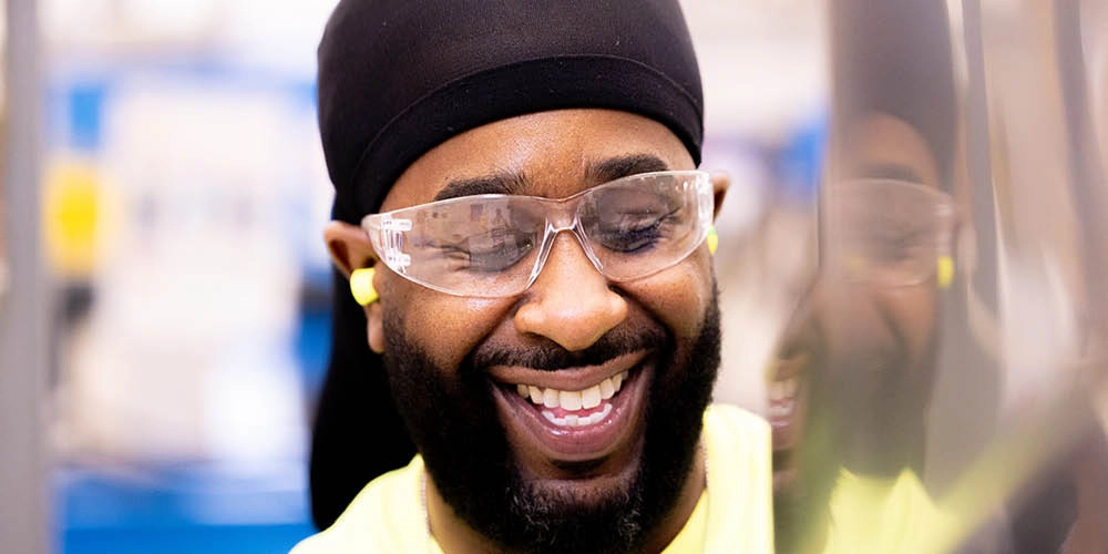 A man wearing safety glasses, earplugs, and a black head covering smiles while working in a brightly lit industrial setting.