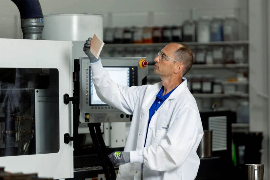 A man in a lab coat and gloves examines a rectangular sample in front of industrial equipment in a laboratory setting.