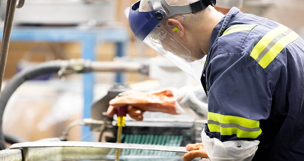 A worker wearing safety gear operates machinery in an industrial setting, with protective gloves, face shield, and earplugs visible.