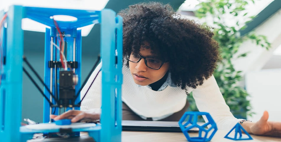 A person adjusts a blue 3D printer on a table, with geometric shapes and a laptop nearby.