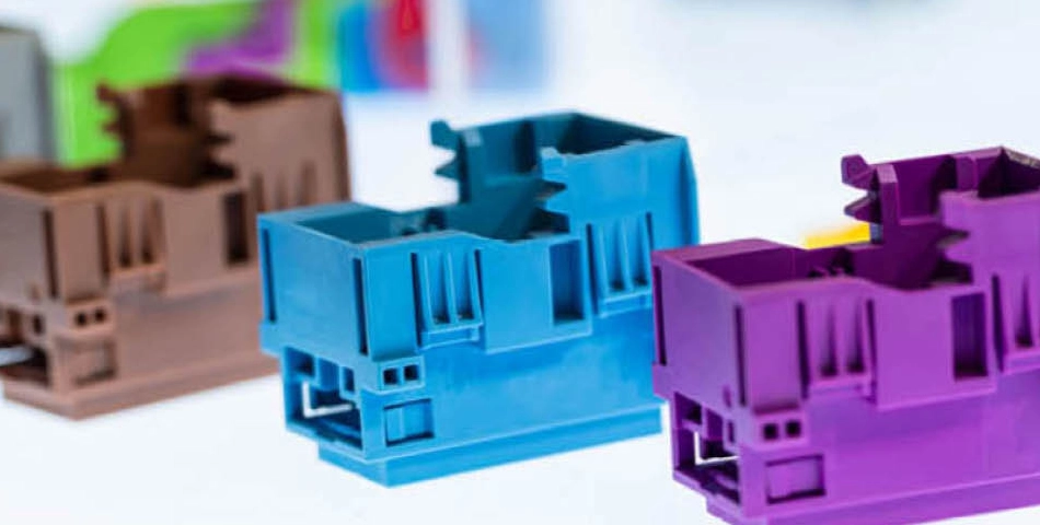 Three plastic electrical housings in brown, blue, and purple are arranged in a row on a white surface, with a blurred colorful background.