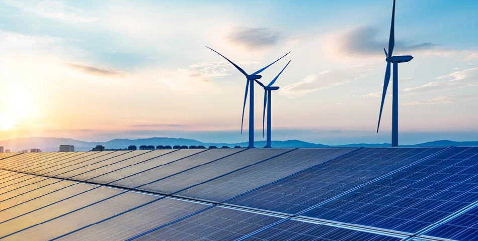 Solar panels and wind turbines stand in a landscape with a mountain range in the background under a partly cloudy sky at sunset.