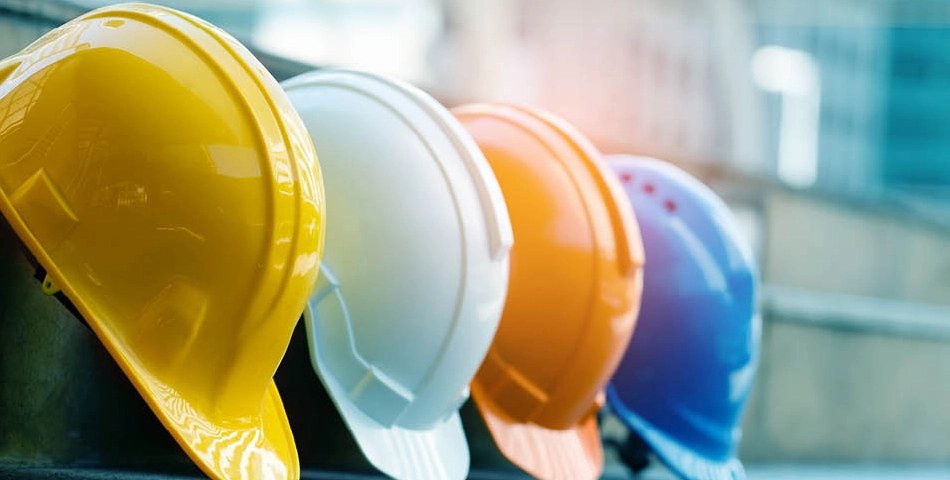Four construction helmets—yellow, white, orange, and blue—are lined up in a row on a concrete surface with a blurred background.