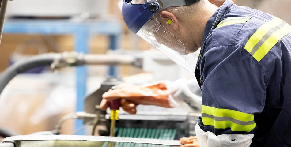 A worker wearing safety gear and a face shield operates machinery in an industrial setting.
