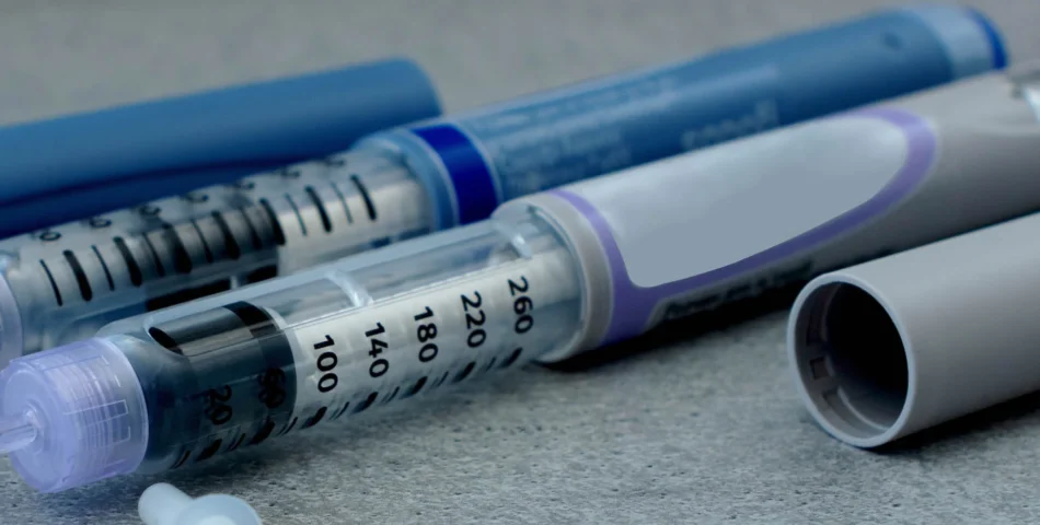 Close-up of insulin pens and their caps on a grey surface. The pens have clear windows showing dosage markings and internal measurements.