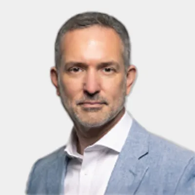 A man with short gray hair and a beard wearing a light blue blazer and white shirt, posing against a plain light background.