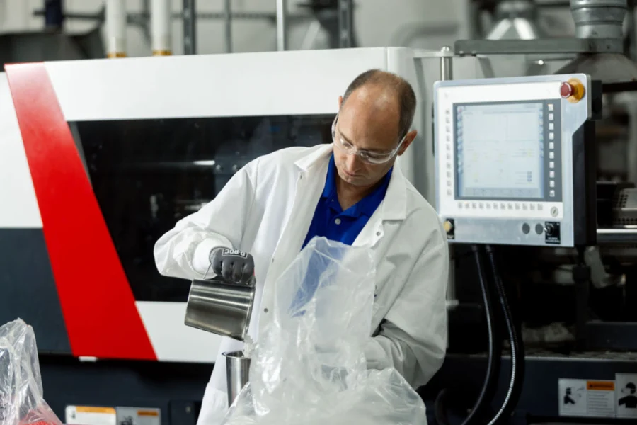 A person in a lab coat and safety glasses pours a substance from a metal container into a plastic bag in an industrial laboratory setting.
