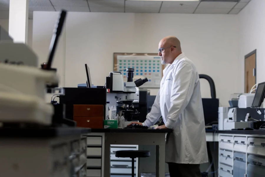 A scientist in a white lab coat stands at a workstation in a laboratory, using a computer and microscope.