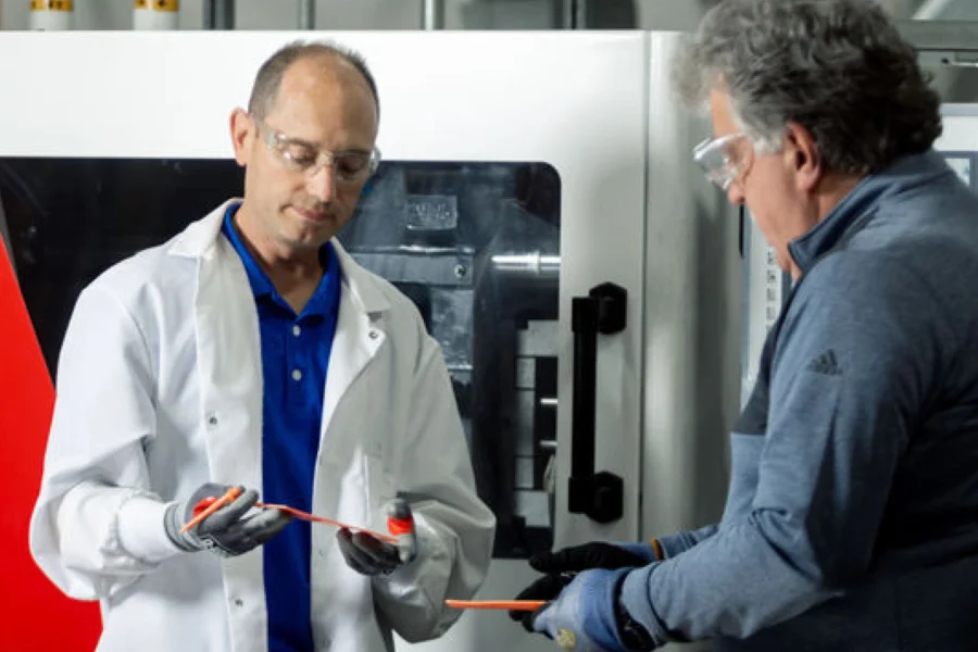 Two men wearing protective eyewear and gloves examine orange plastic parts in an industrial setting.