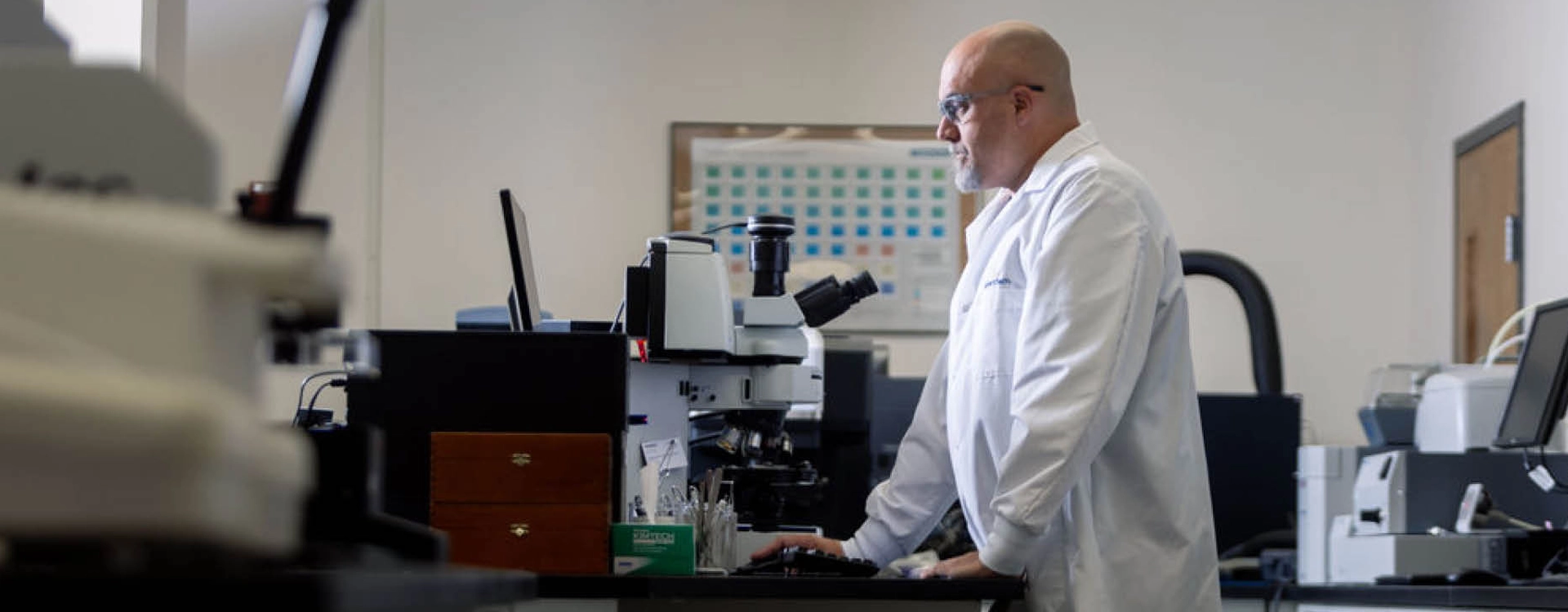A scientist in a white lab coat works at a computer station in a laboratory, with lab equipment and a microscope nearby.