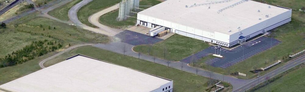 Aerial view of two large industrial buildings with adjacent parking lots, surrounded by grass and a few trees, connected by paved roads.