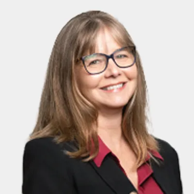 Woman with straight, light brown hair and glasses, wearing a dark blazer over a maroon blouse, smiling at the camera against a plain light background.