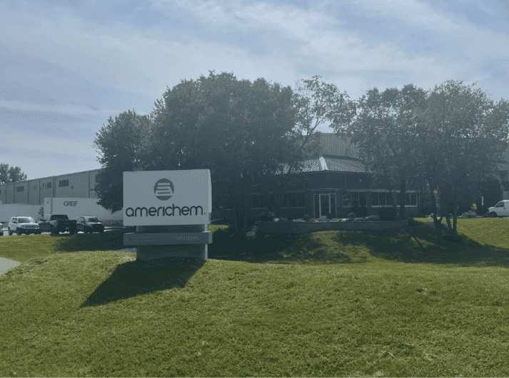 A sign for Americhem stands in front of a commercial building surrounded by trees and grass under a partly cloudy sky.