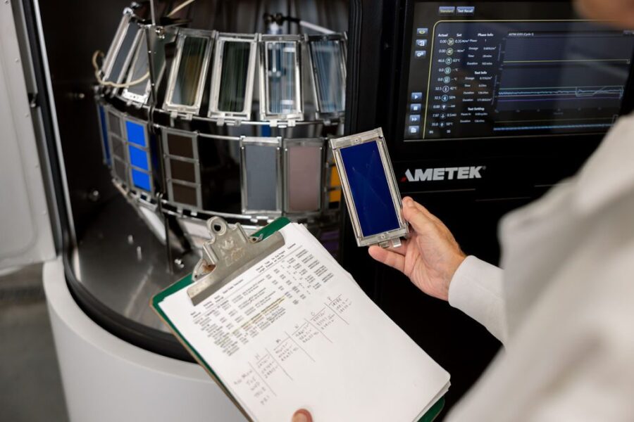 A person in a lab coat holds a blue sample panel near a machine displaying panels; a clipboard with printed data is held in the other hand.