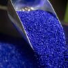 A metal scoop pours small, bright blue plastic pellets onto a pile of similar pellets.
