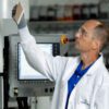 A scientist in a lab coat and safety glasses examines a rectangular sample in front of laboratory equipment.