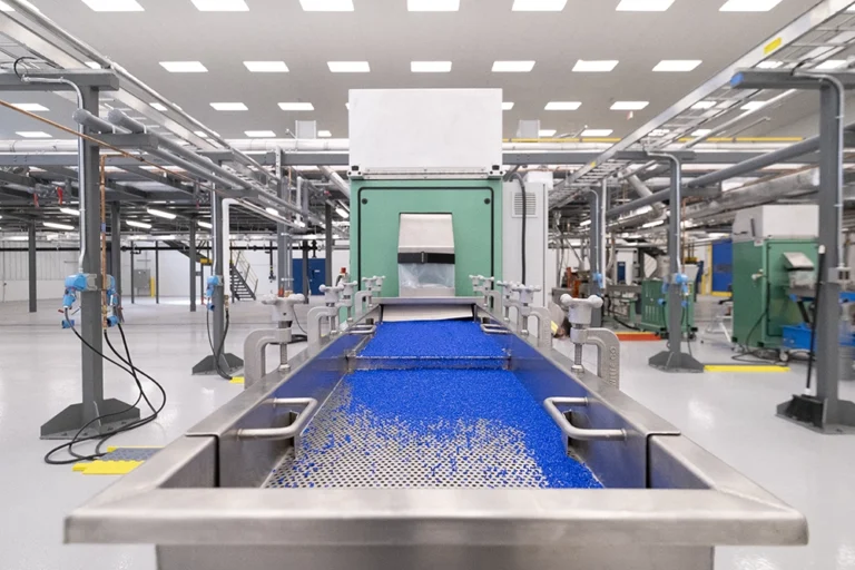 A factory interior with a stainless steel conveyor transporting blue plastic pellets toward a large green industrial machine under bright overhead lights.
