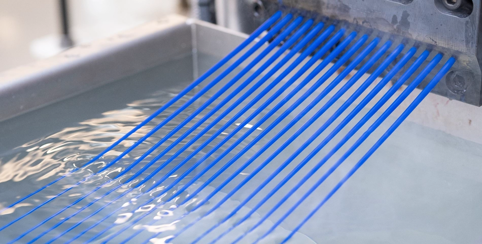 Several blue plastic filaments are being extruded from a machine into a water bath for cooling.