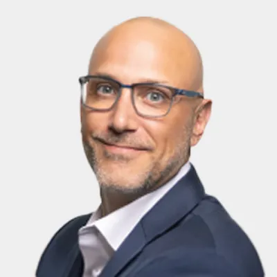 Bald man with glasses wearing a dark suit and white shirt, smiling slightly, posed against a plain light background.