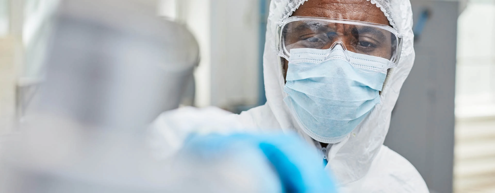A scientist wearing protective clothing, a face mask, and goggles works in a laboratory setting.