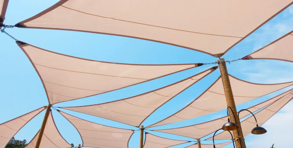 Light brown fabric shade sails are stretched between poles, forming a geometric canopy under a blue sky. Two outdoor lamps are visible below the canopy.
