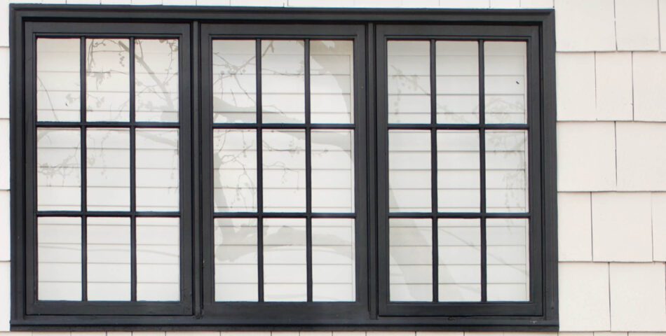 A three-panel window with black grid frames, set in a white shingled exterior wall, with white blinds partially drawn behind the glass.