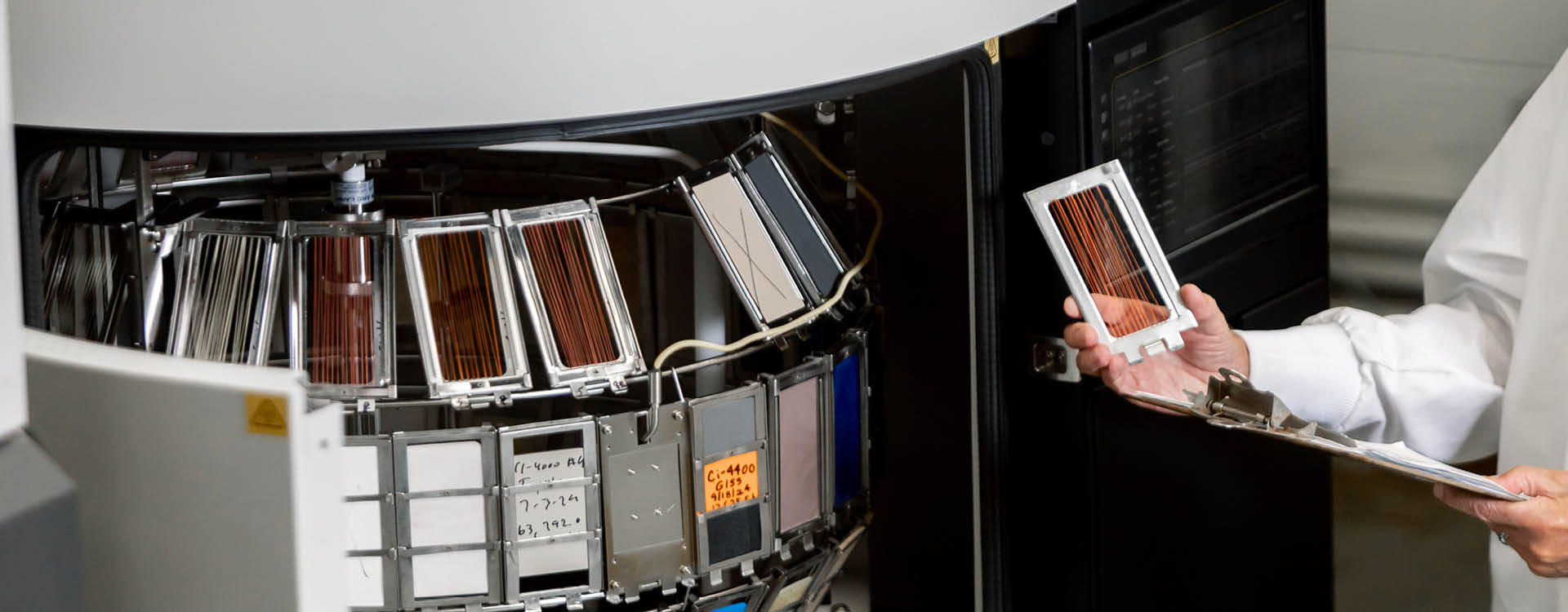 A person holds a solar panel sample near a machine containing multiple solar panel samples arranged in slots; a clipboard is also visible.