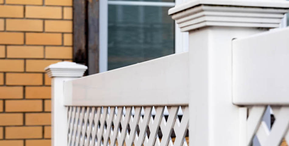 Close-up of a white lattice fence with square posts in front of a window and a yellow brick wall.