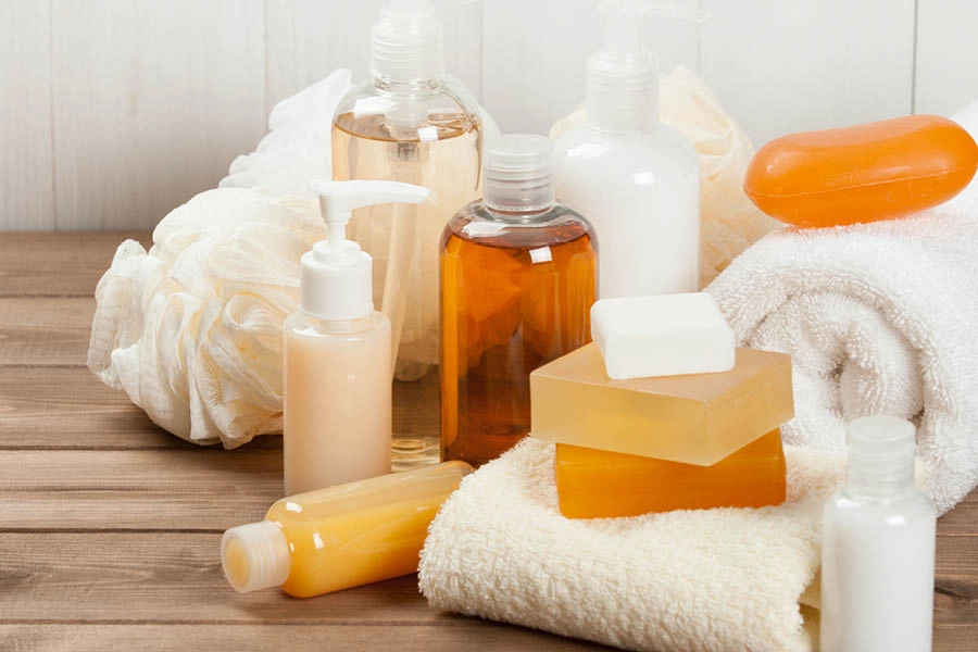 Assorted toiletry items including liquid bottles, bars of soap, a white towel, and a bath sponge arranged on a wooden surface.