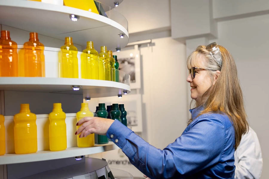 A woman in a blue shirt selects a yellow bottle from a curved shelf displaying bottles in various shades of yellow and green.