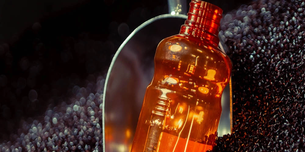 A close-up of an amber-colored plastic bottle with a red cap, surrounded by dark granules and a reflective surface in the background.