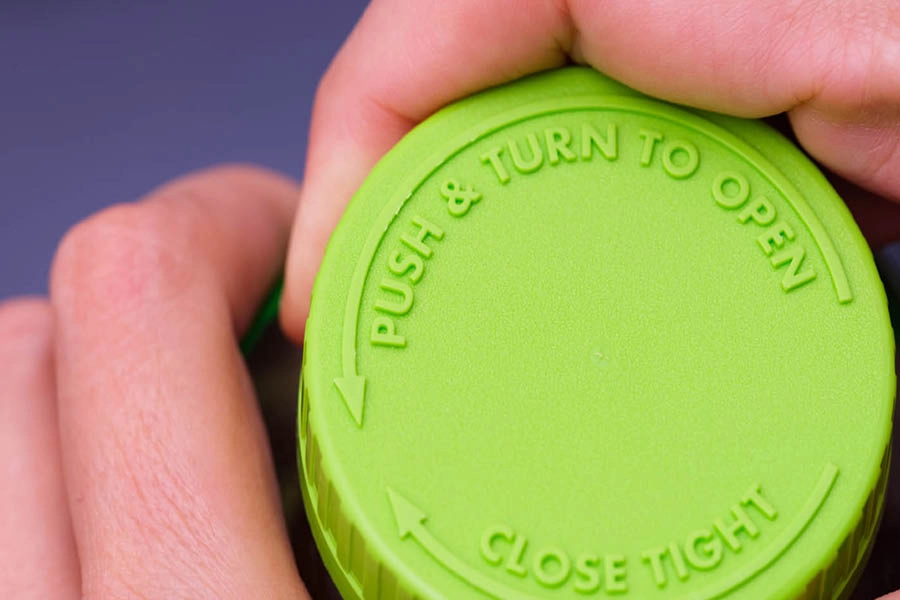 A close-up of a green bottle cap with the instructions "PUSH & TURN TO OPEN" and "CLOSE TIGHT" embossed on the top, held by two hands.