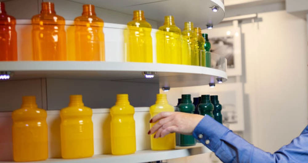 A person’s hand reaches toward colorful bottles arranged by shade on curved white shelves, ranging from orange to yellow to green.