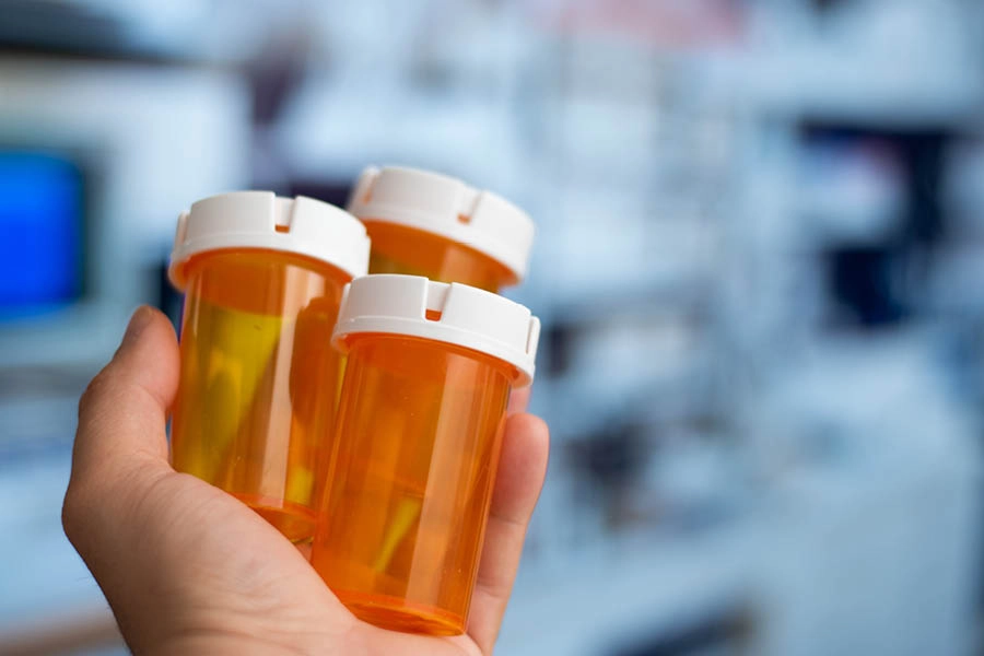 A hand holds three empty orange prescription pill bottles with white caps against a blurred background.