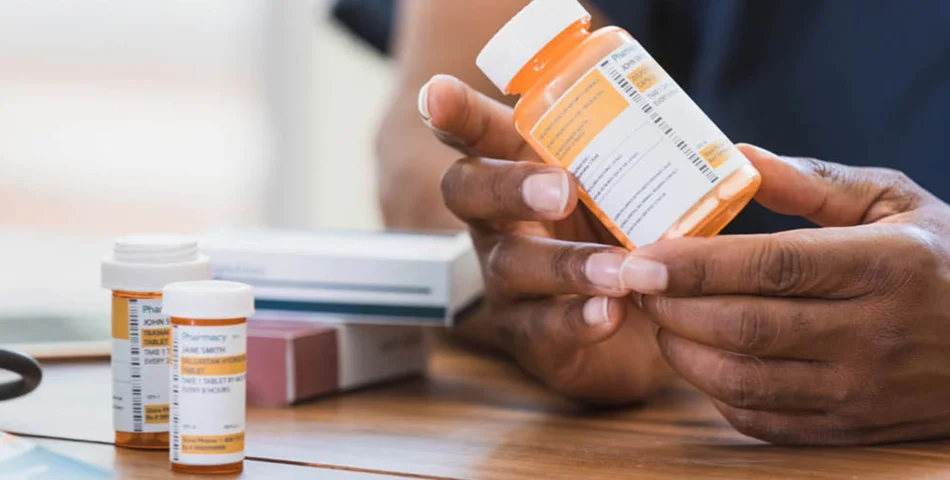 A person holds a prescription pill bottle while other medication bottles and boxes are on a wooden table.