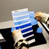 A person compares a blue plastic sample from Americhem with a Pantone color chart featuring various shades of blue on a table.