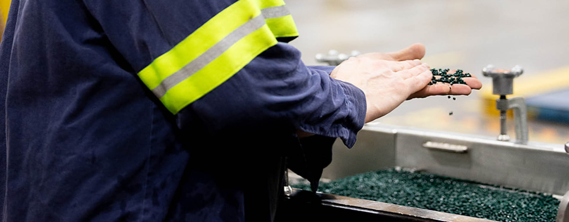 A worker in a blue uniform with a yellow reflective stripe examines small black plastic pellets by hand at an industrial workstation.
