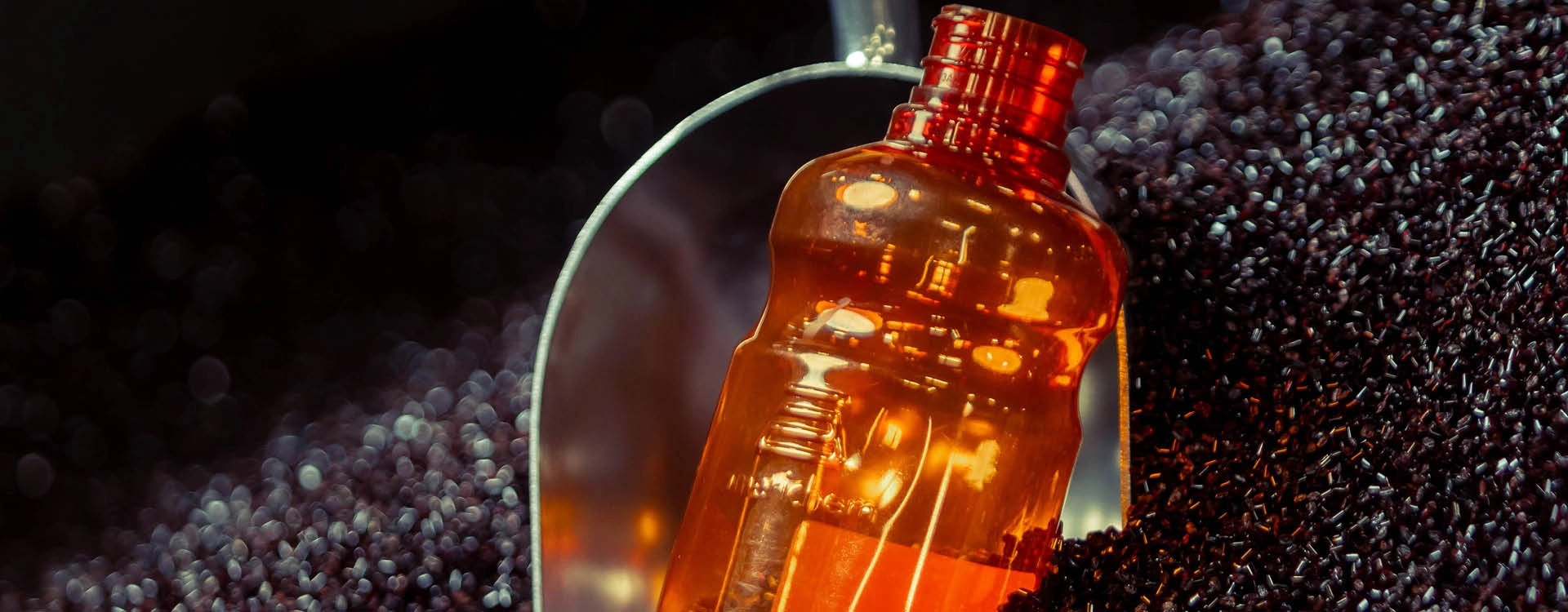 A transparent orange plastic bottle is lying on black plastic pellets with a metal scoop partially visible in the background.