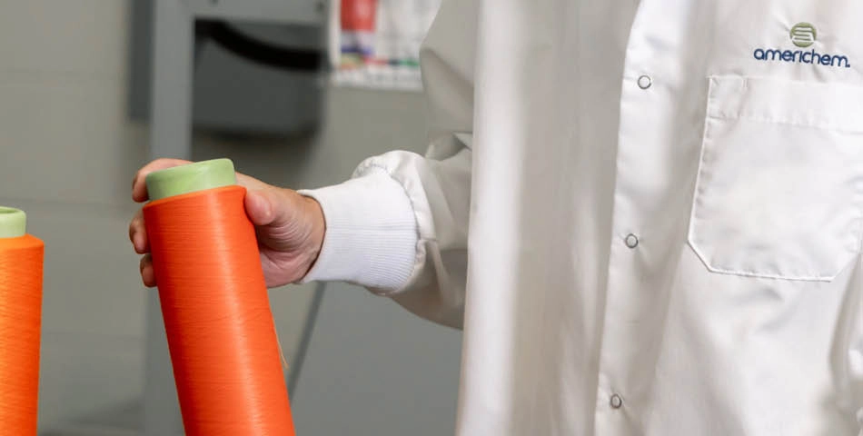 Person wearing a white lab coat holding a spool of orange thread or yarn in a laboratory or manufacturing setting.