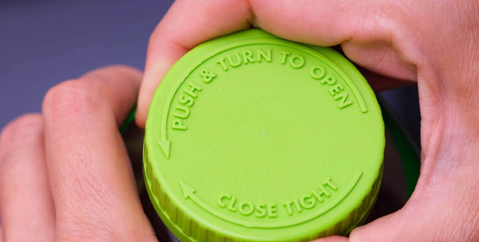 Close-up of hands gripping a green child-proof bottle cap with embossed instructions: "PUSH & TURN TO OPEN" and "CLOSE TIGHT" with directional arrows.