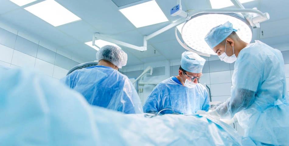 Three surgeons in blue scrubs and masks perform an operation in a brightly lit, sterile operating room.