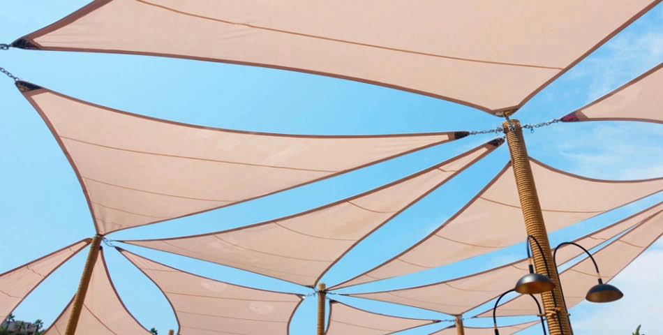 Beige fabric shade sails are stretched between poles, providing cover under a blue sky. Multiple poles and lamps are visible beneath the triangular sails.
