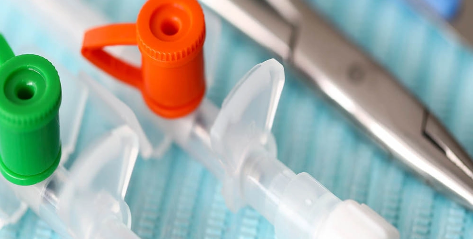 Close-up of medical equipment including IV cannulas with green and orange caps and a pair of metal forceps on a blue textured surface.