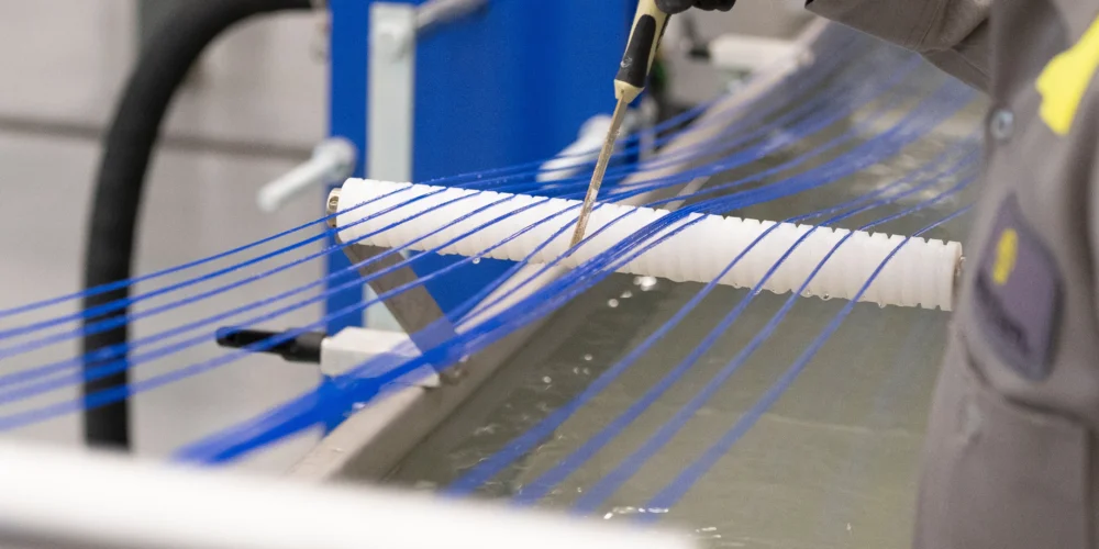 A person uses a tool to guide multiple blue strings over a white cylindrical rod above a tank of liquid in an industrial setting.