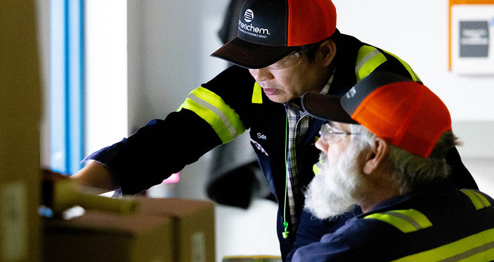Two workers wearing safety jackets and orange caps review documents together at a worksite.