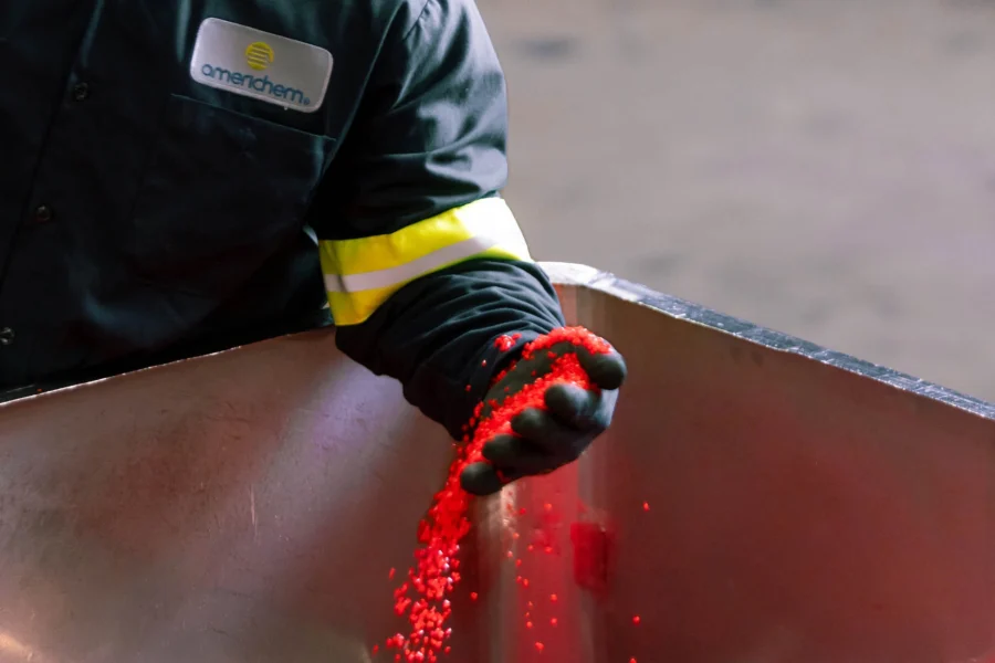 A worker in protective clothing pours red plastic pellets from a container, with the Americhem logo visible on the uniform.