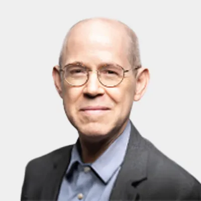 Bald man with glasses wearing a dark blazer and light blue shirt, smiling slightly, on a plain light background.
