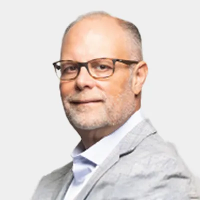 A middle-aged man with glasses, short gray hair, and a beard wearing a light gray suit jacket and white shirt, posing against a plain light background.